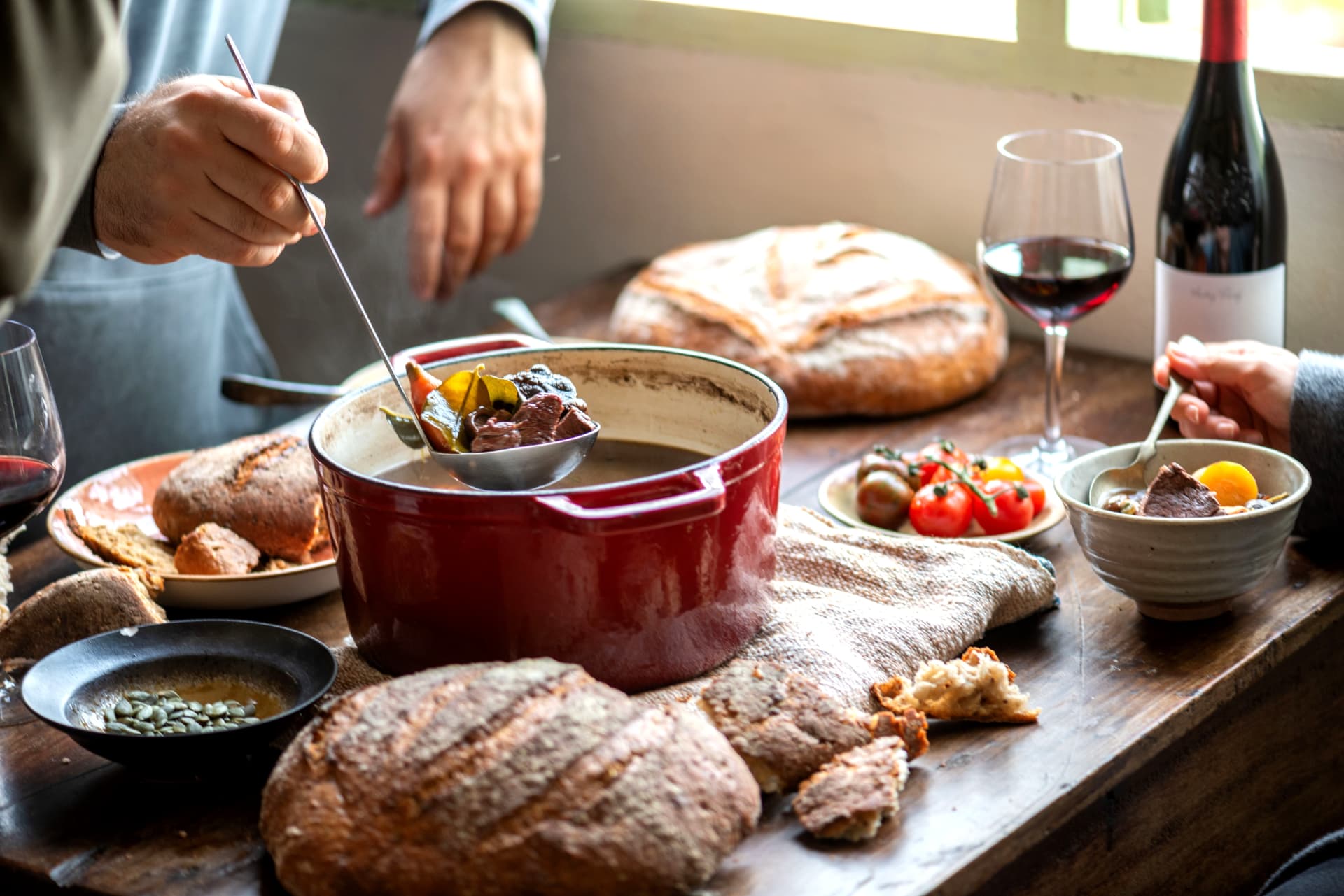 Hands serving a braise from a red Dutch oven with bread and wine on the table